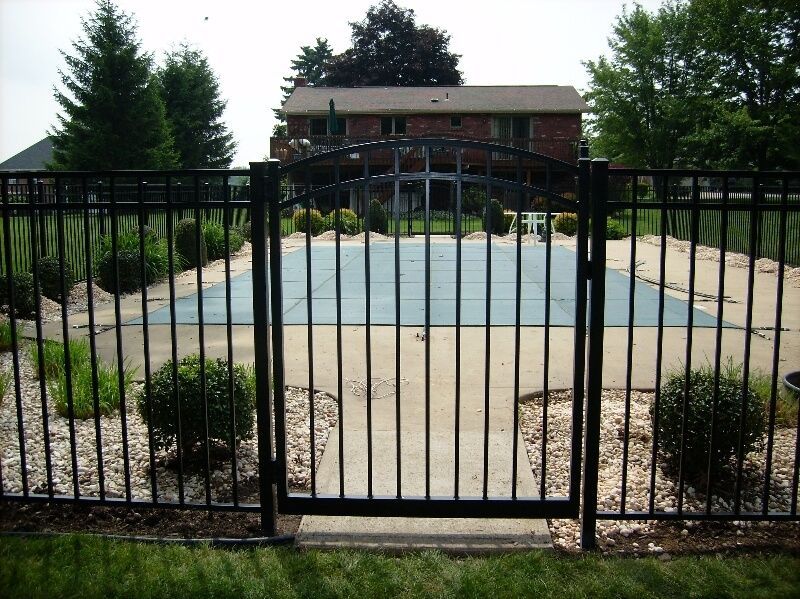 Black metal fence in front of an empty outdoor swimming pool with a brick house and trees beyond