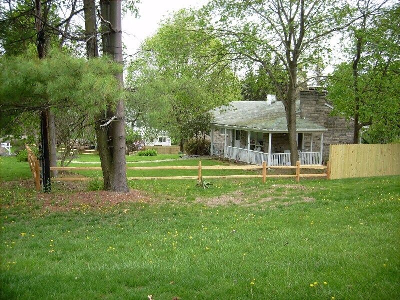 Grassy yard with trees and a small house with a porch, seen behind a low fence