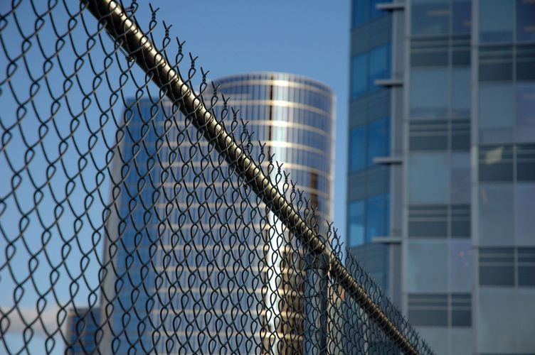 Chain-link fence in foreground with modern office buildings against a blue sky