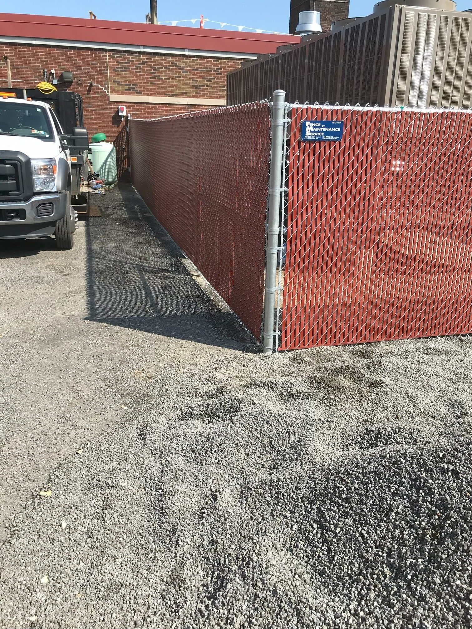 Alley beside red chain-link fence and brick building, with a parked truck on gravel.