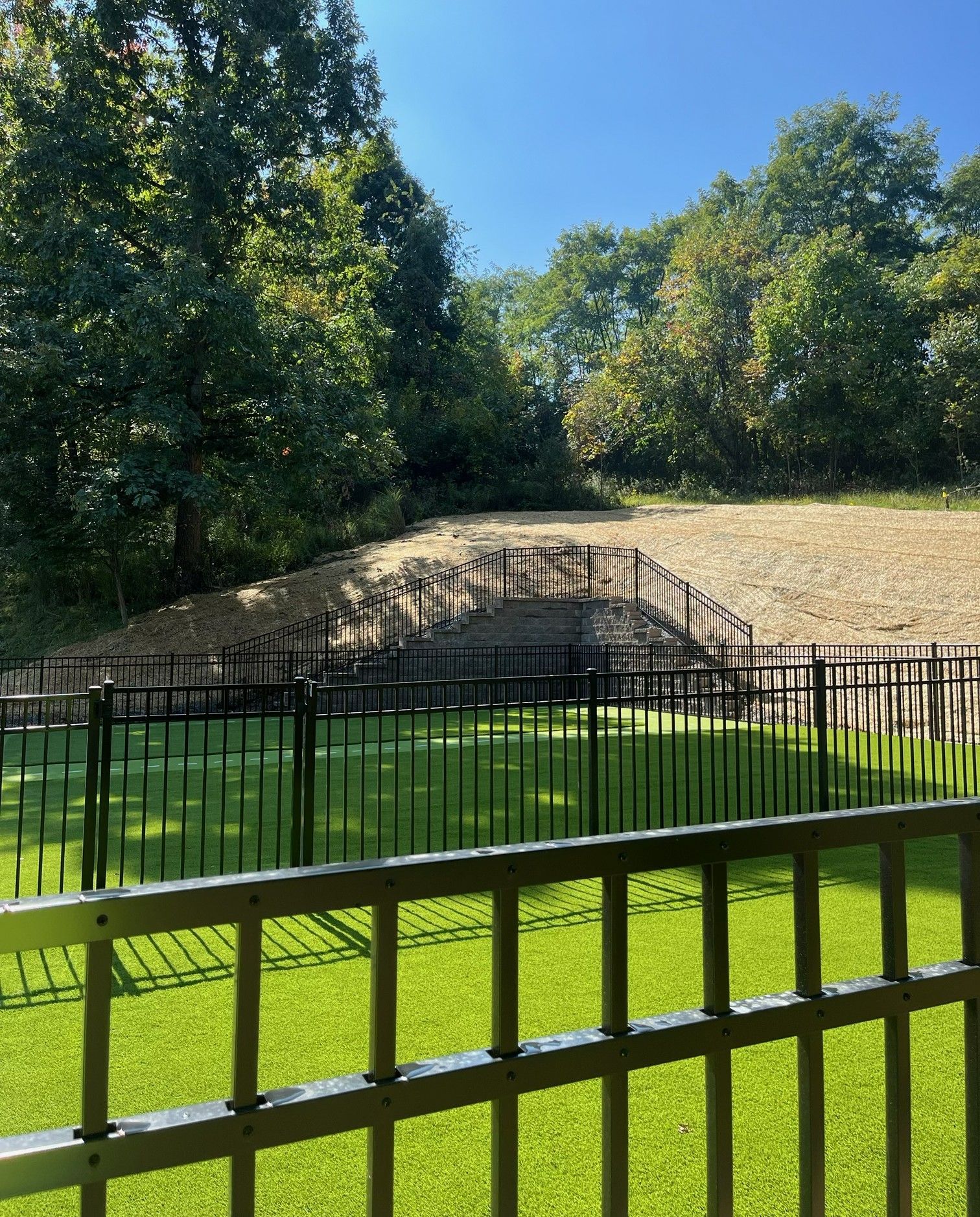 Grassy enclosure with a black fence, stone mound, and trees under a clear blue sky