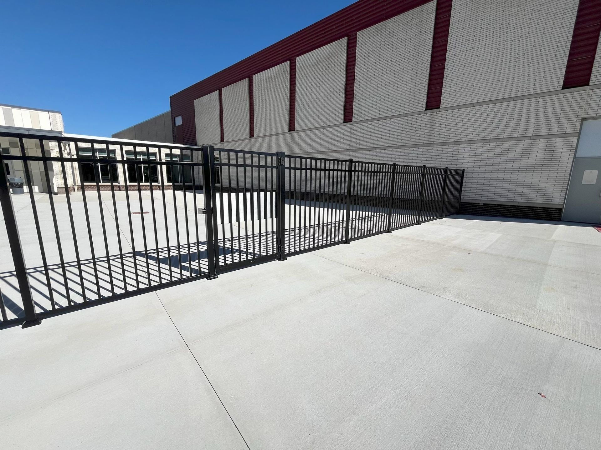 Empty concrete courtyard with black metal fencing beside a red-and-white building under a clear blue sky