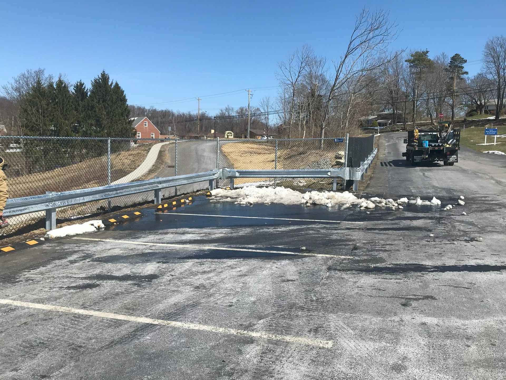 Snow-covered parking lot with a metal guardrail, a road, and a parked vehicle under a clear blue sky