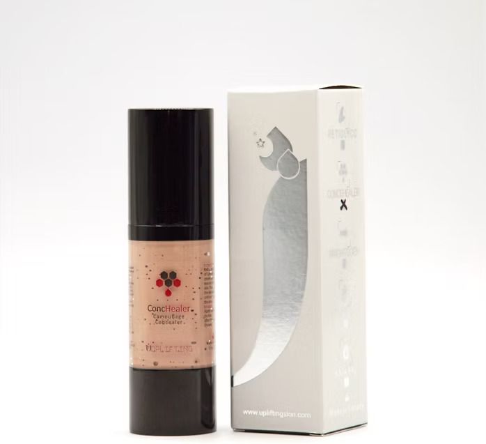 A makeup foundation bottle and box. The bottle is black with a label. The box is white with a silver logo.