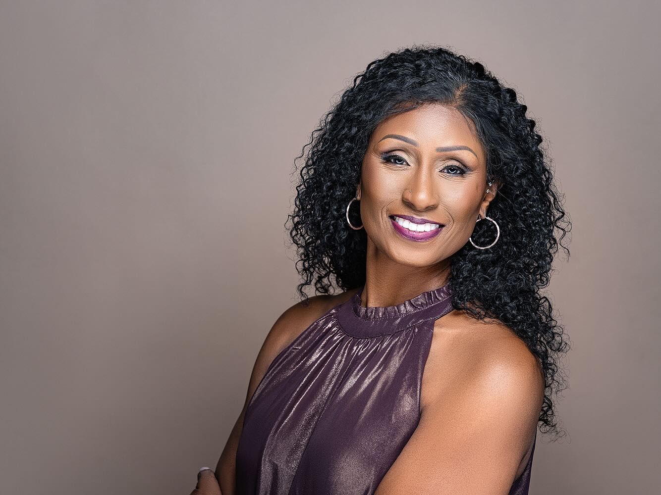 Woman with curly hair in a purple dress smiling, arms crossed, on a gray background.