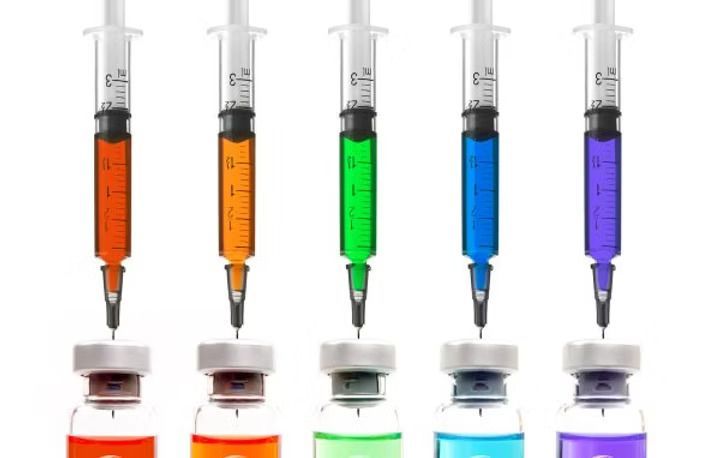 Five syringes filled with different colored liquids poised above vials, white background.