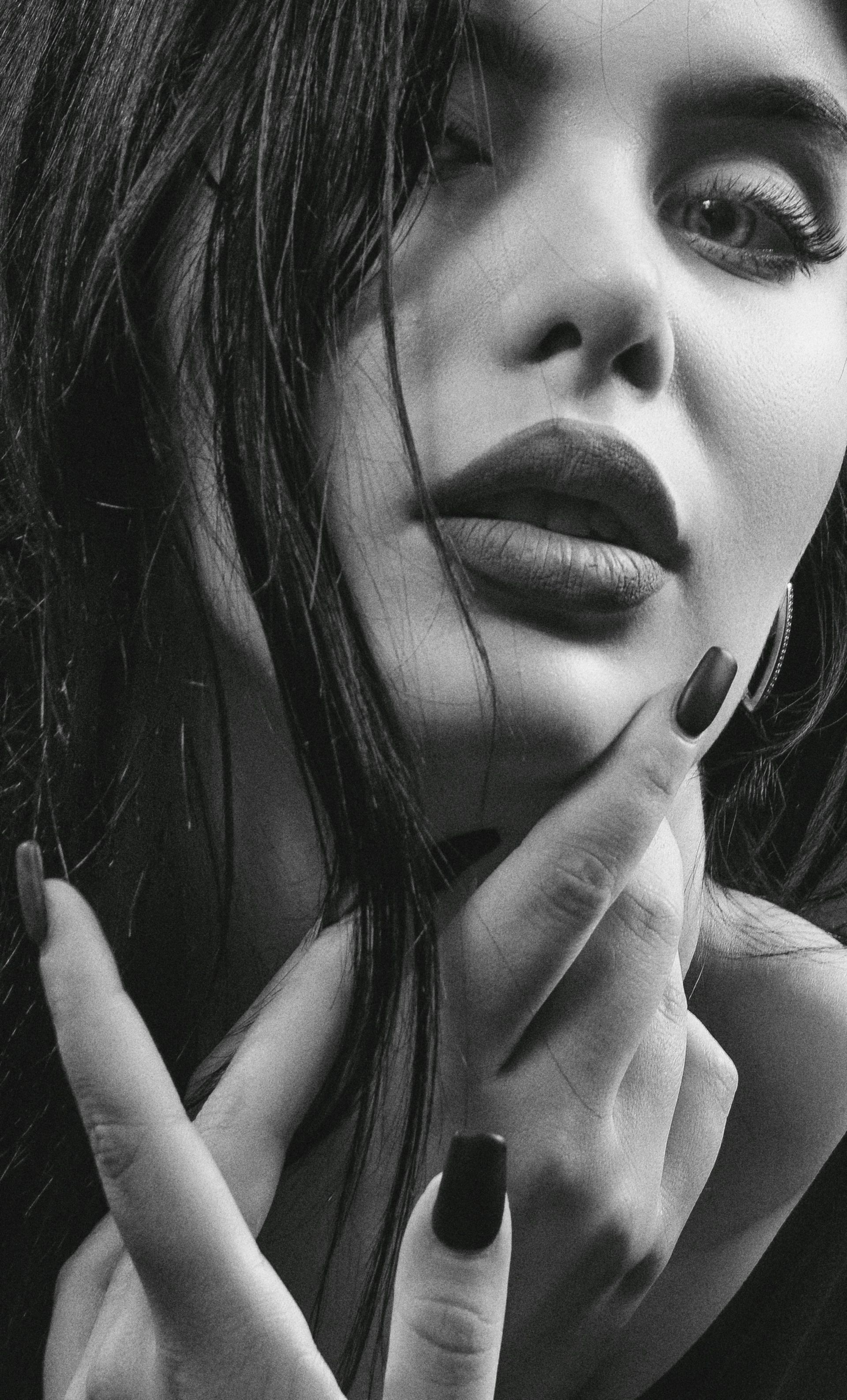 A close-up, black-and-white portrait of a person with dark hair, dark lipstick, and dark nail polish posing near their face.