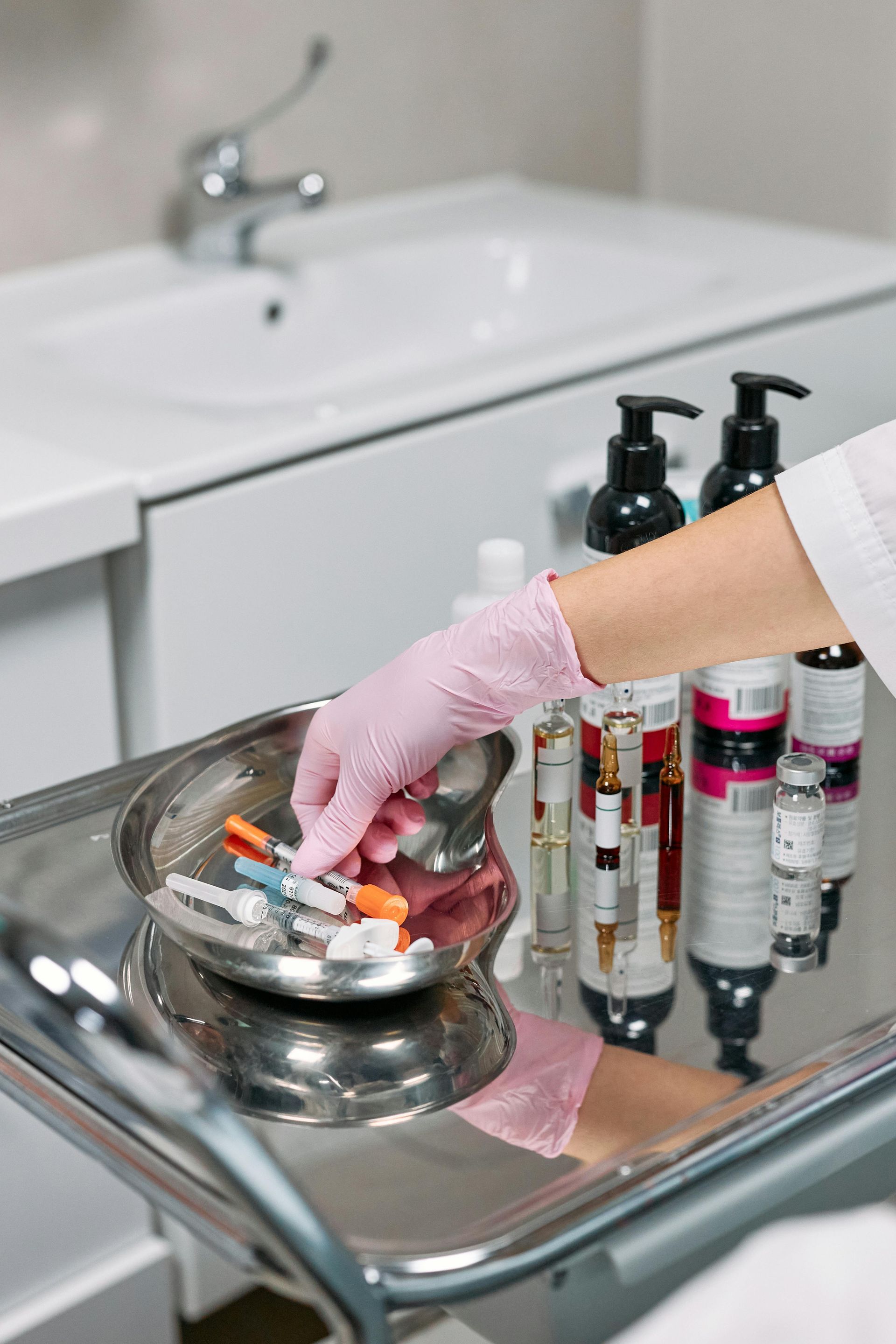 Person in pink gloves reaching for syringes in a metal tray, with cosmetic products and a sink in the background.