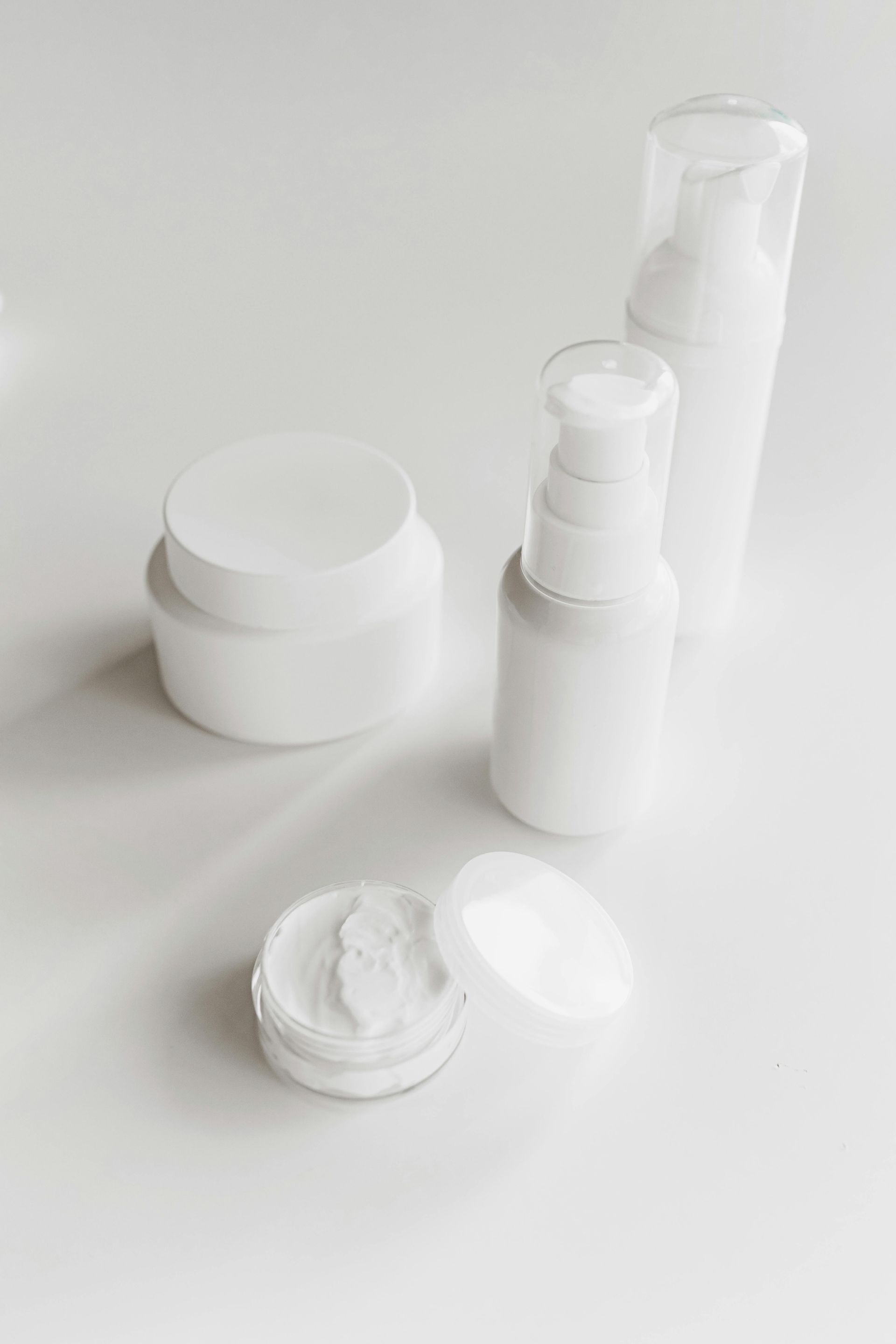 White skincare products on a white surface: a jar with cream, bottles and a container with a lid.