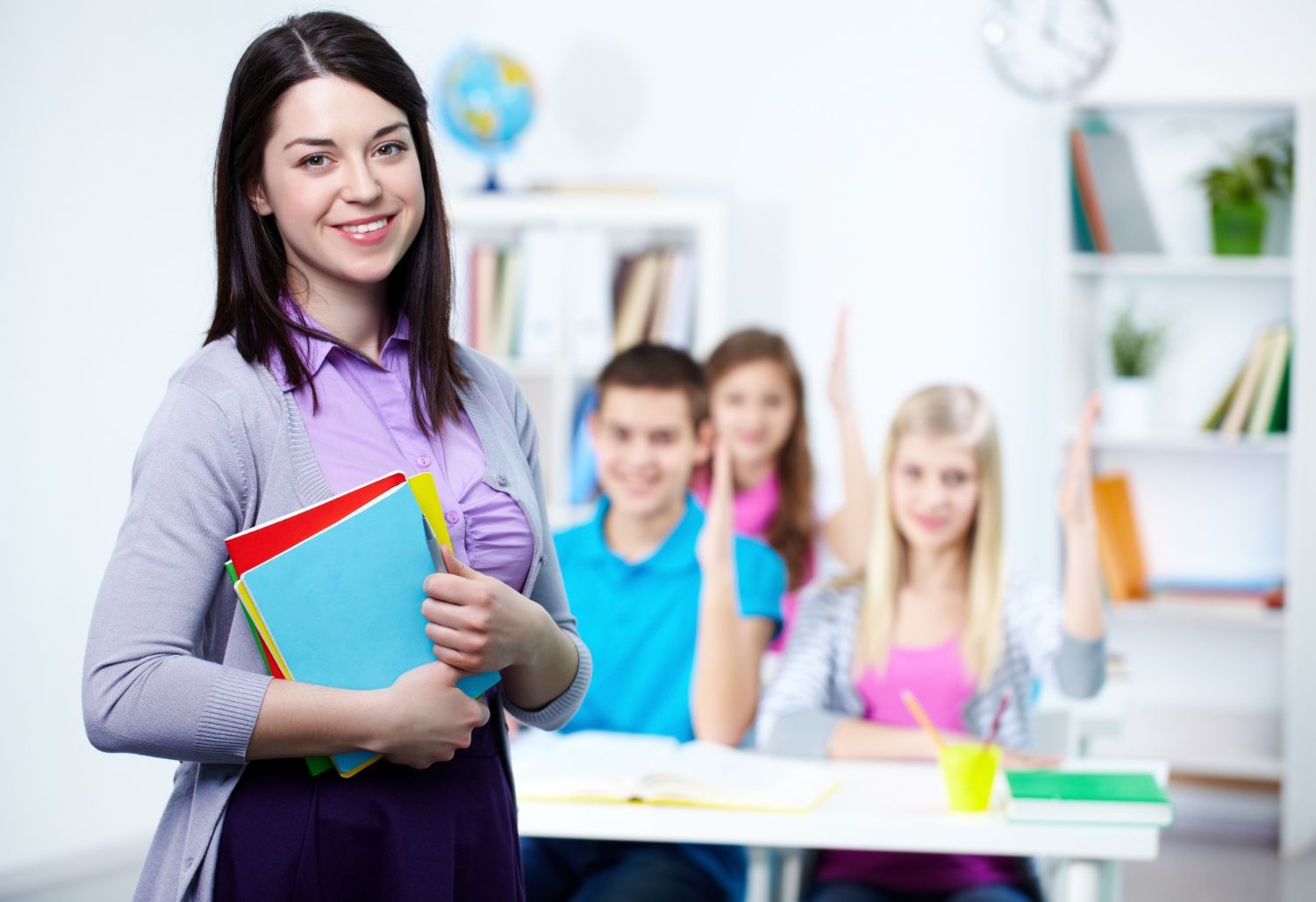 A teacher is standing in front of her students in a classroom holding books.