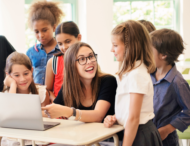 A group of children are listening to their female teacher as she explains something on her laptop.