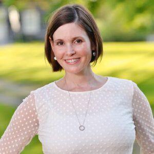 A woman wearing a white polka dot shirt and a necklace is smiling for the camera.