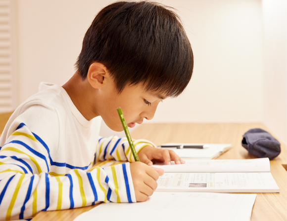 Boy in striped shirt writing at a desk, focused on a book and paper.
