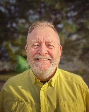 A man with a beard is wearing a yellow shirt and smiling.