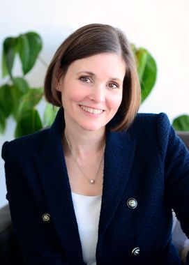 Woman with short brown hair smiles, wearing a navy blazer, white top, and necklace. Blurred green plants in background.