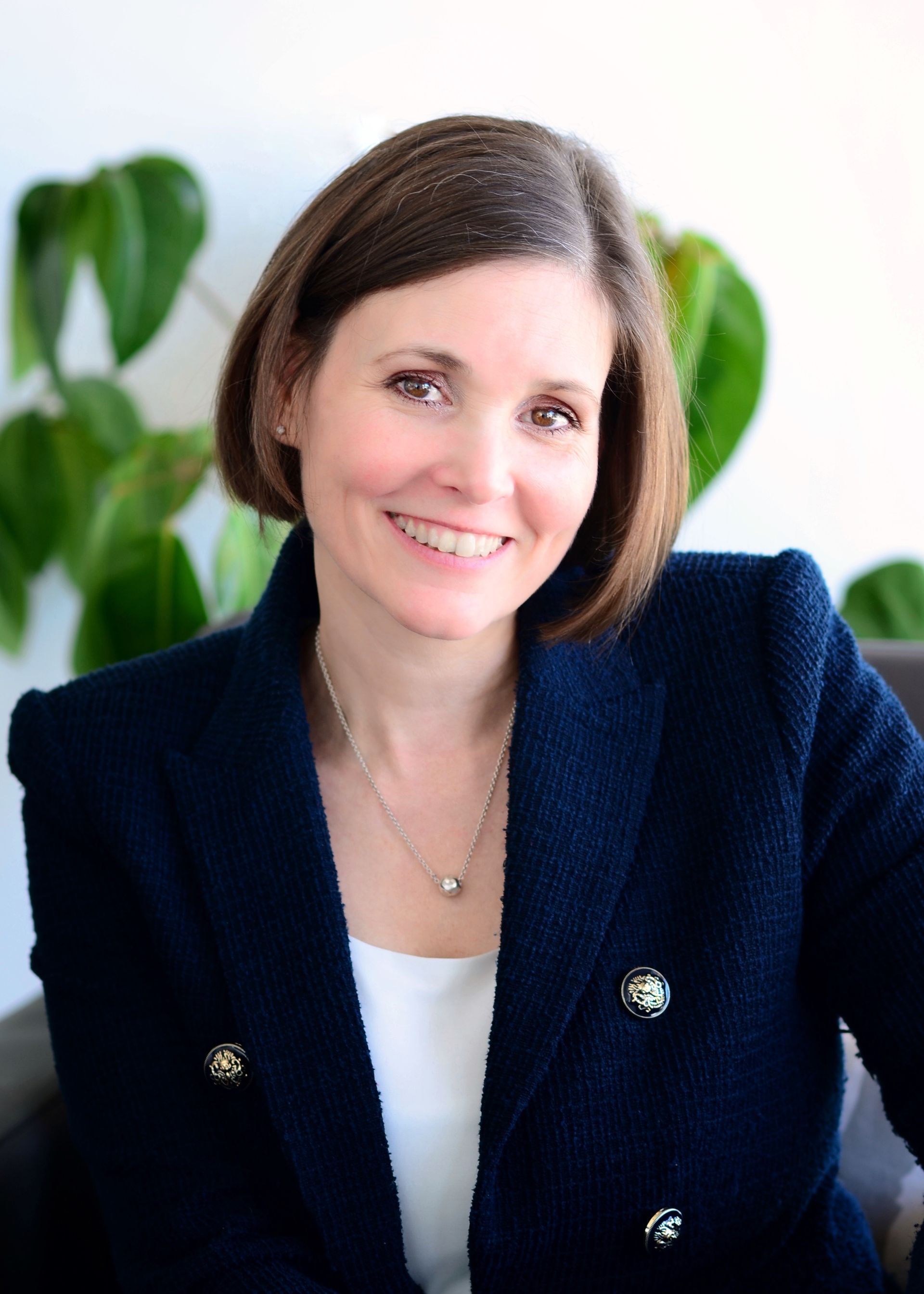 Woman with short brown hair smiles, wearing a navy blazer, white top, and necklace. Blurred green plants in background.