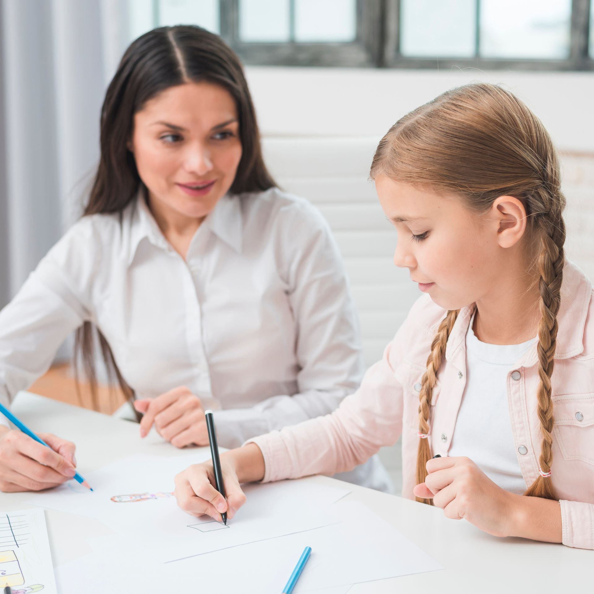 A woman and a little girl are sitting at a table writing on a piece of paper.