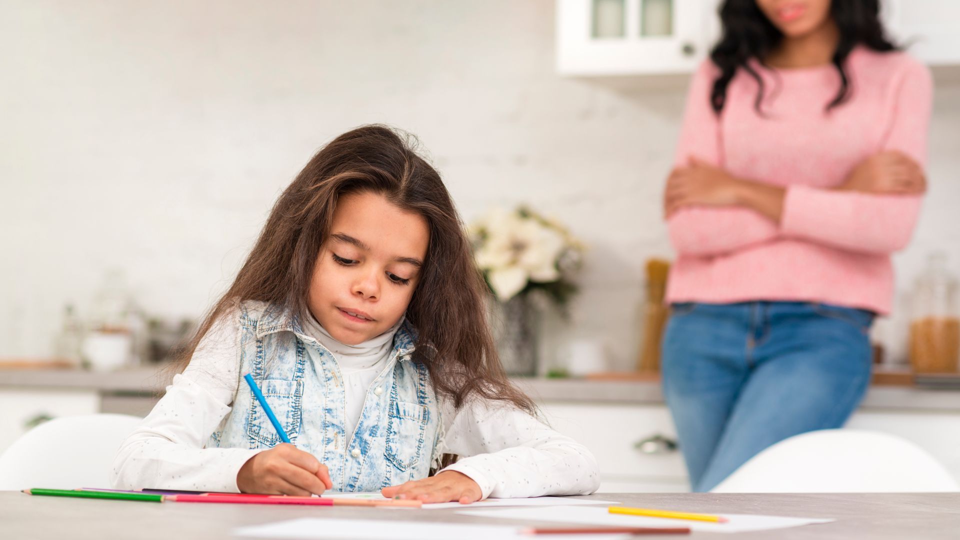 A little girl is sitting at a table drawing with a pencil while a woman stands behind her.