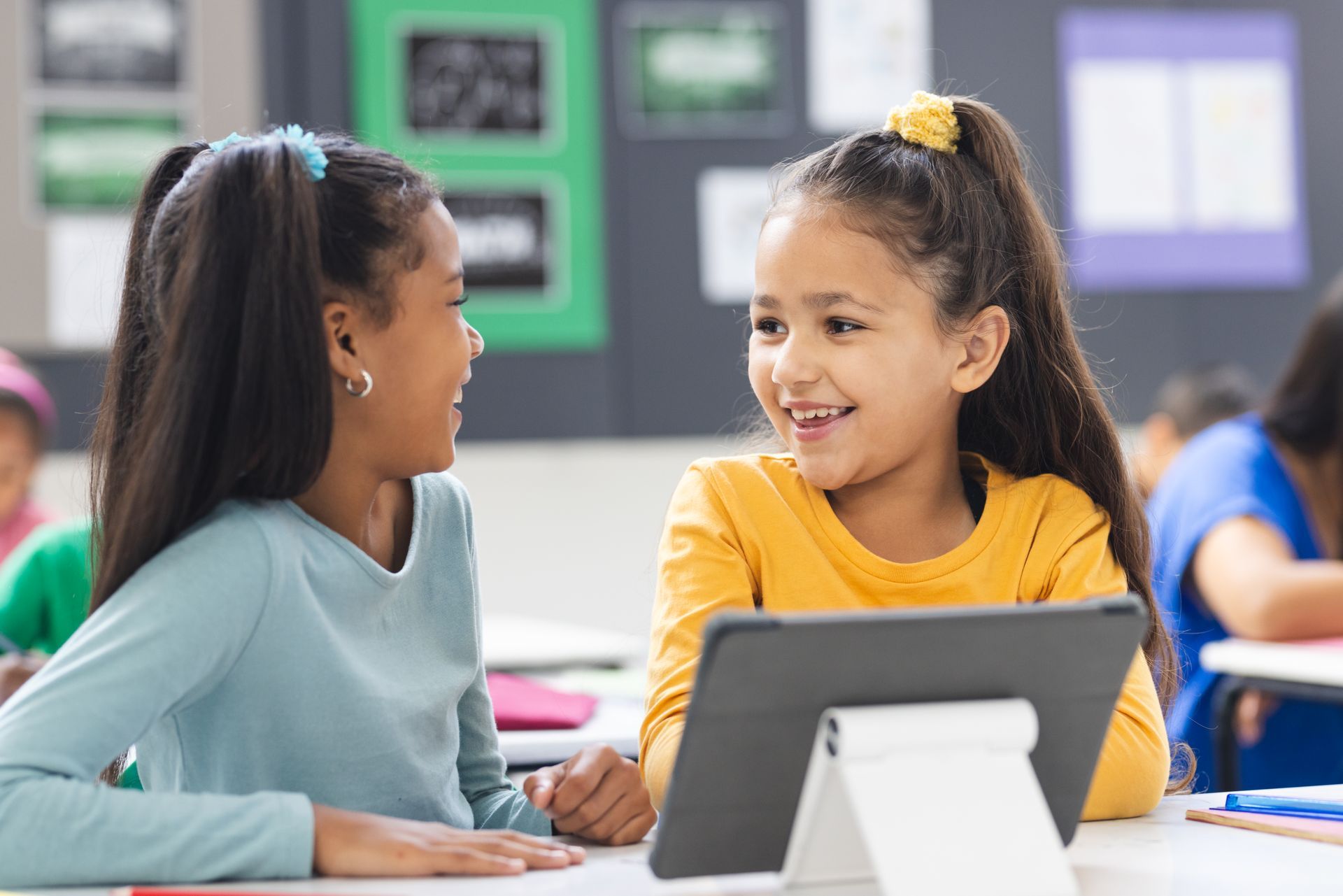 Two young girls are sitting at a desk in a classroom looking at a tablet.