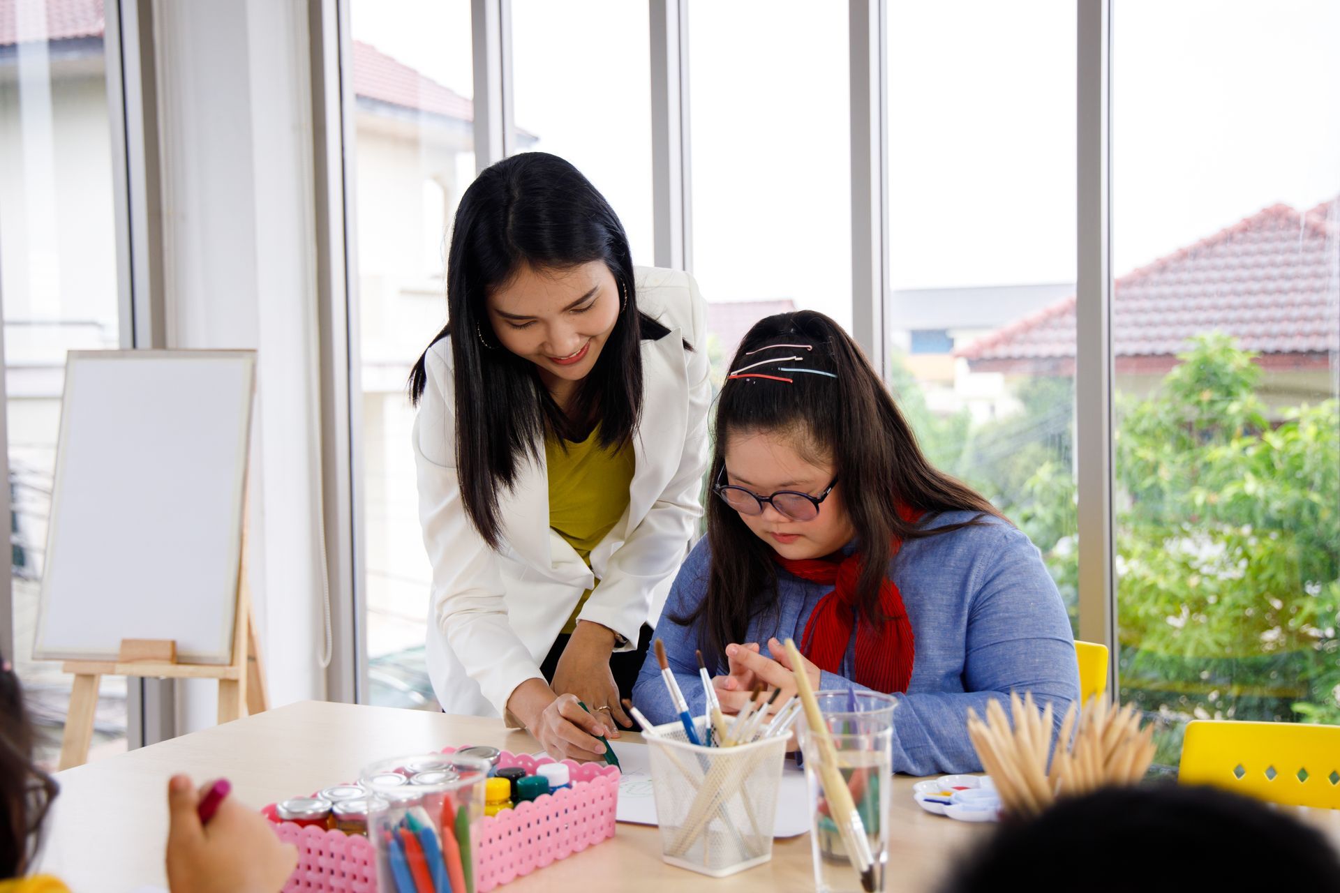 A woman is standing next to a girl sitting at a table.