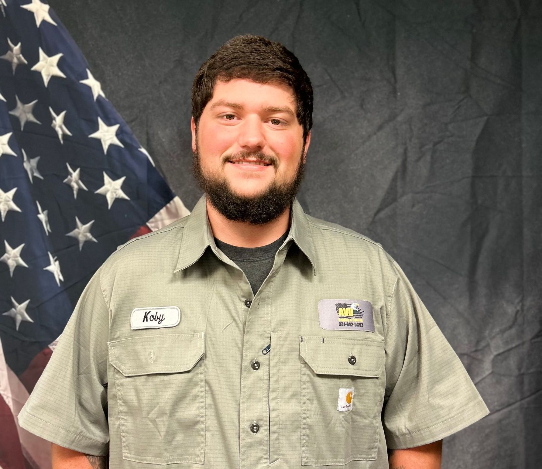 A man with a beard is standing in front of an american flag.