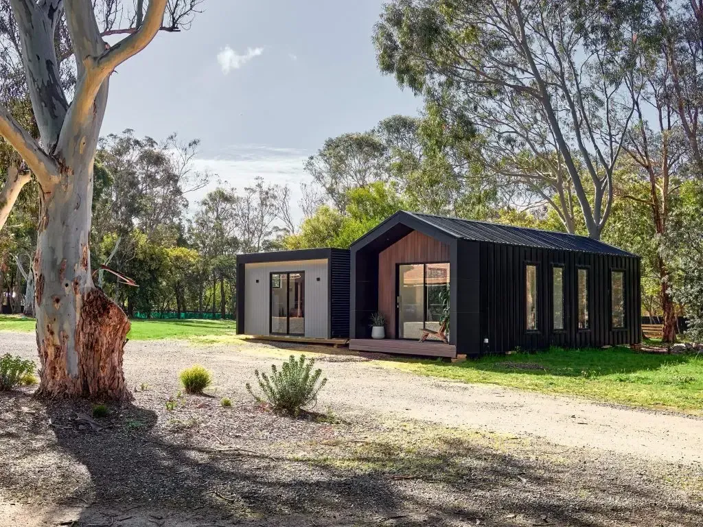 Modern black and grey house with large windows on a gravel driveway surrounded by trees and greenery.