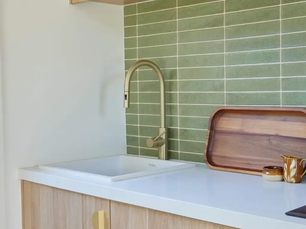 Kitchen sink with gold faucet against green tiled wall; wooden countertop, tray, and cabinets.