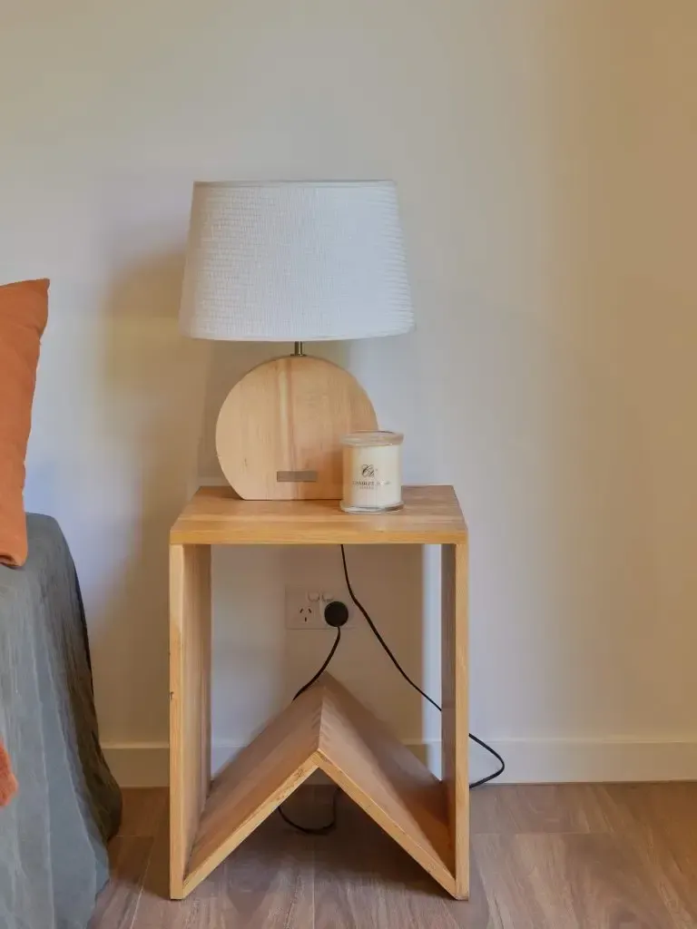 Wooden side table with a lamp, candle, and orange pillow against a white wall.
