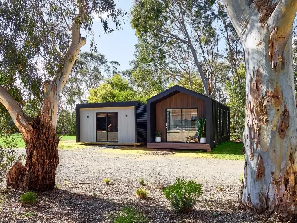 Two modern cabins in a grassy area, with trees framing the view. Gray and brown exteriors.