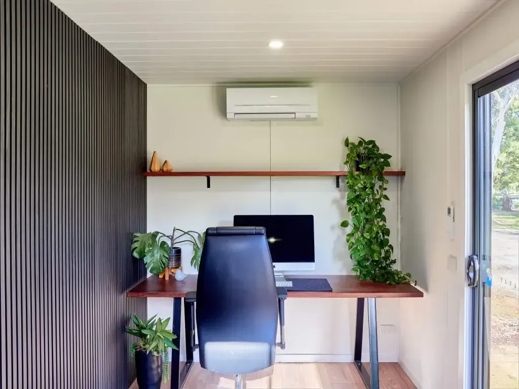 Home office with desk, plants, black chair, white walls, dark wood accents, and air conditioning unit.