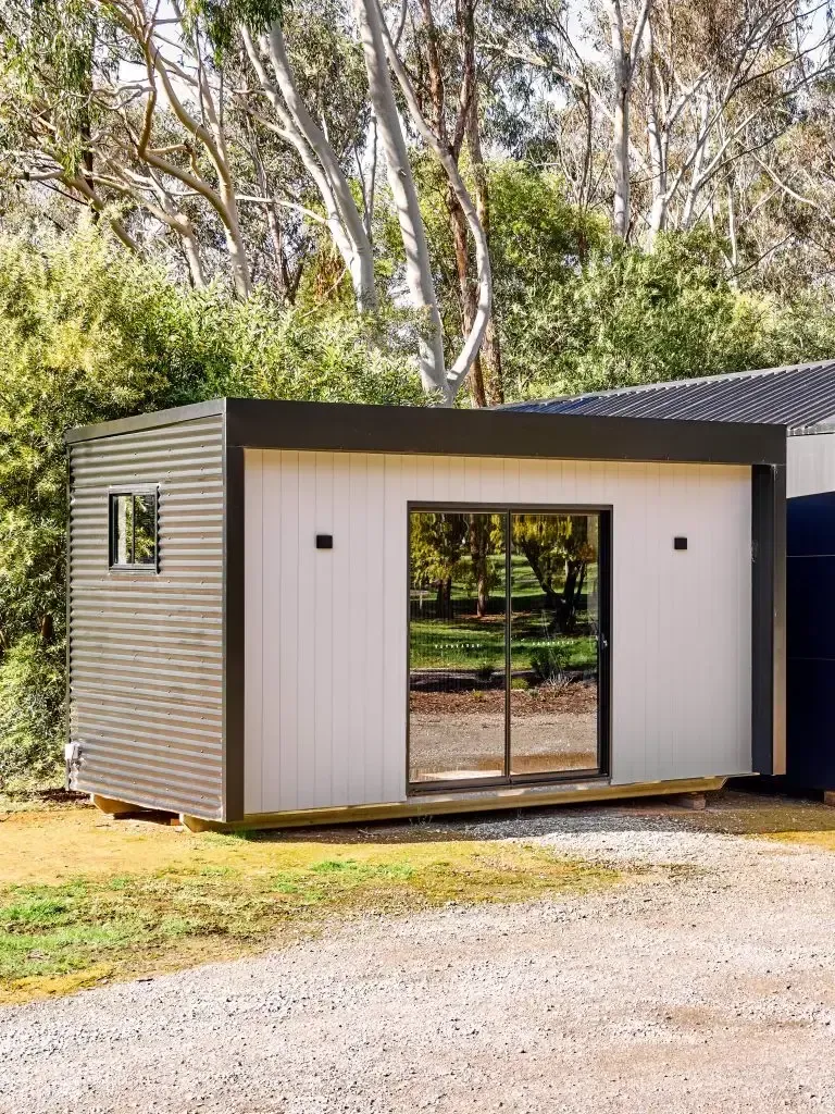 Modern rectangular building with white walls, sliding glass door, and corrugated side, set in a yard.