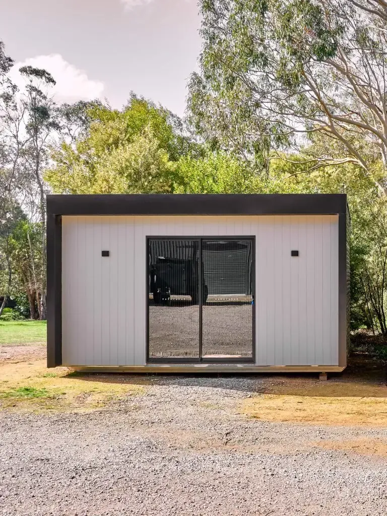 Modern, light gray cabin with dark trim and glass doors, set in a gravel clearing surrounded by trees.