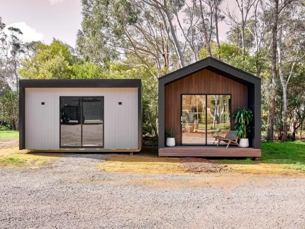 Two modern, small cabins in a gravel yard, with one featuring a sloped roof and wooden siding.