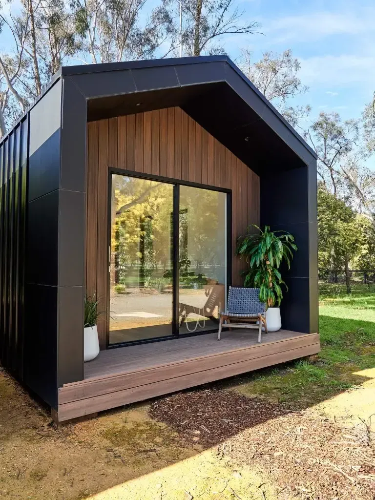 Modern black and brown cabin with glass sliding doors, small deck, and greenery outside.