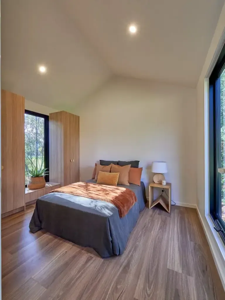 Bedroom with wooden floor, bed with orange and grey bedding, large windows, and light wood storage.