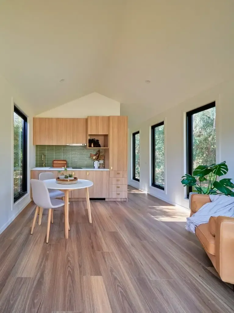 Light-filled interior with kitchen, small round table, and leather sofa by windows overlooking greenery.