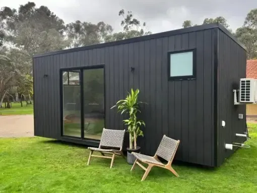 Black tiny home with sliding glass door and small window, two chairs, and a plant on a grassy lawn.