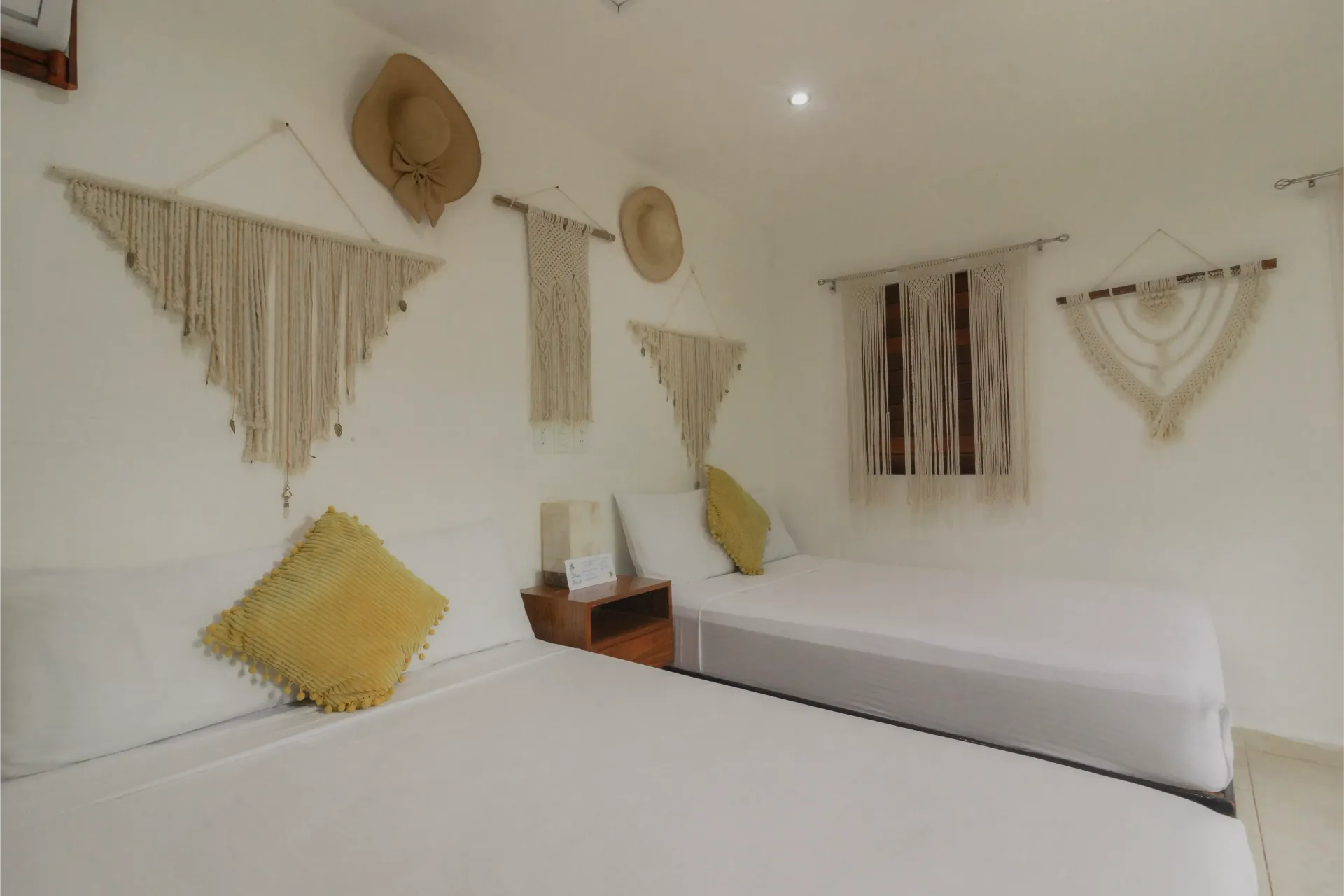 Habitación luminosa y minimalista con paredes blancas, tapices de macramé y una cama con almohadas amarillas y blancas.