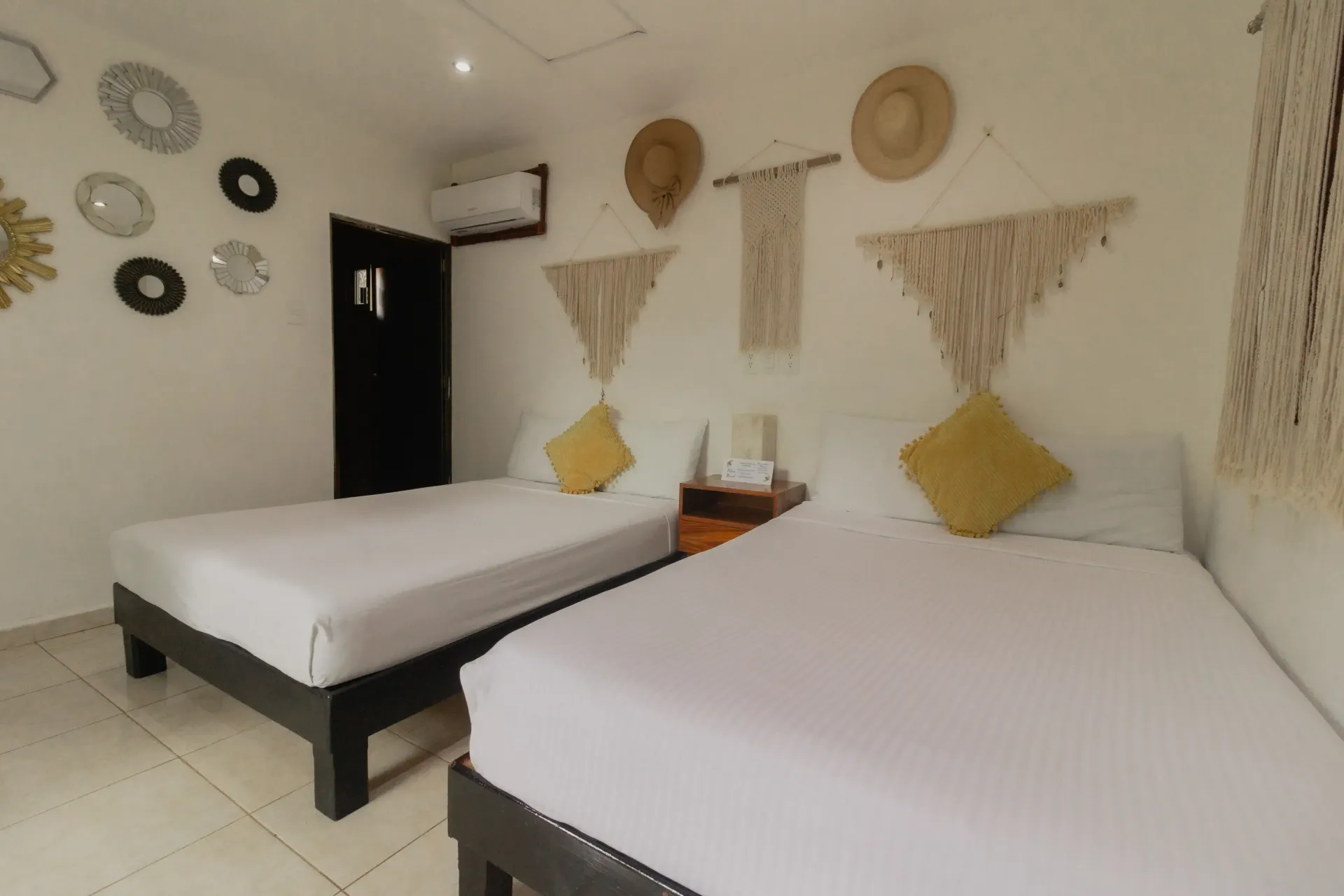 Dormitorio minimalista con dos camas, paredes blancas, decoración de pared tejida y cojines amarillos.