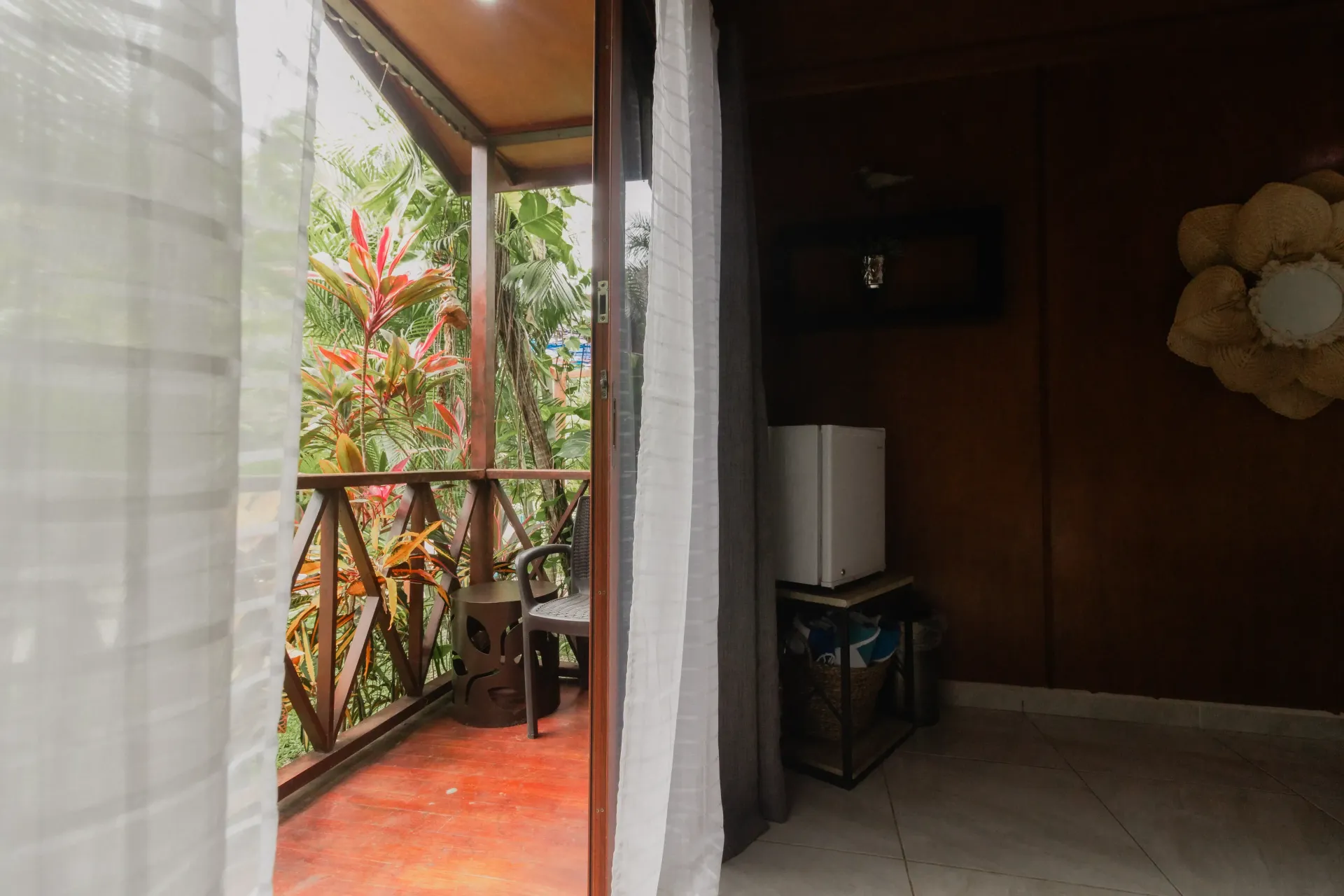 Porche cubierto con barandilla de madera, suelo de baldosas rojas y una puerta interior oscura junto a cortinas blancas.