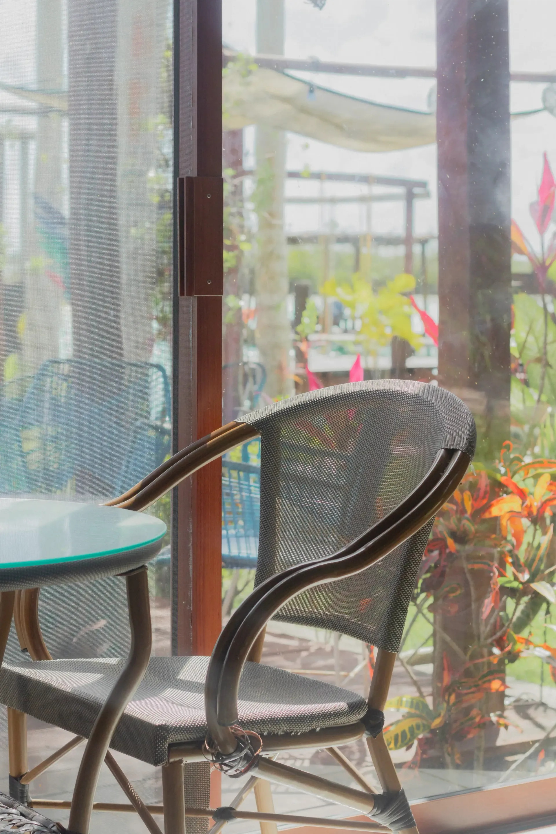 Silla de mimbre y mesa turquesa junto a una ventana luminosa con plantas de jardín en el exterior.