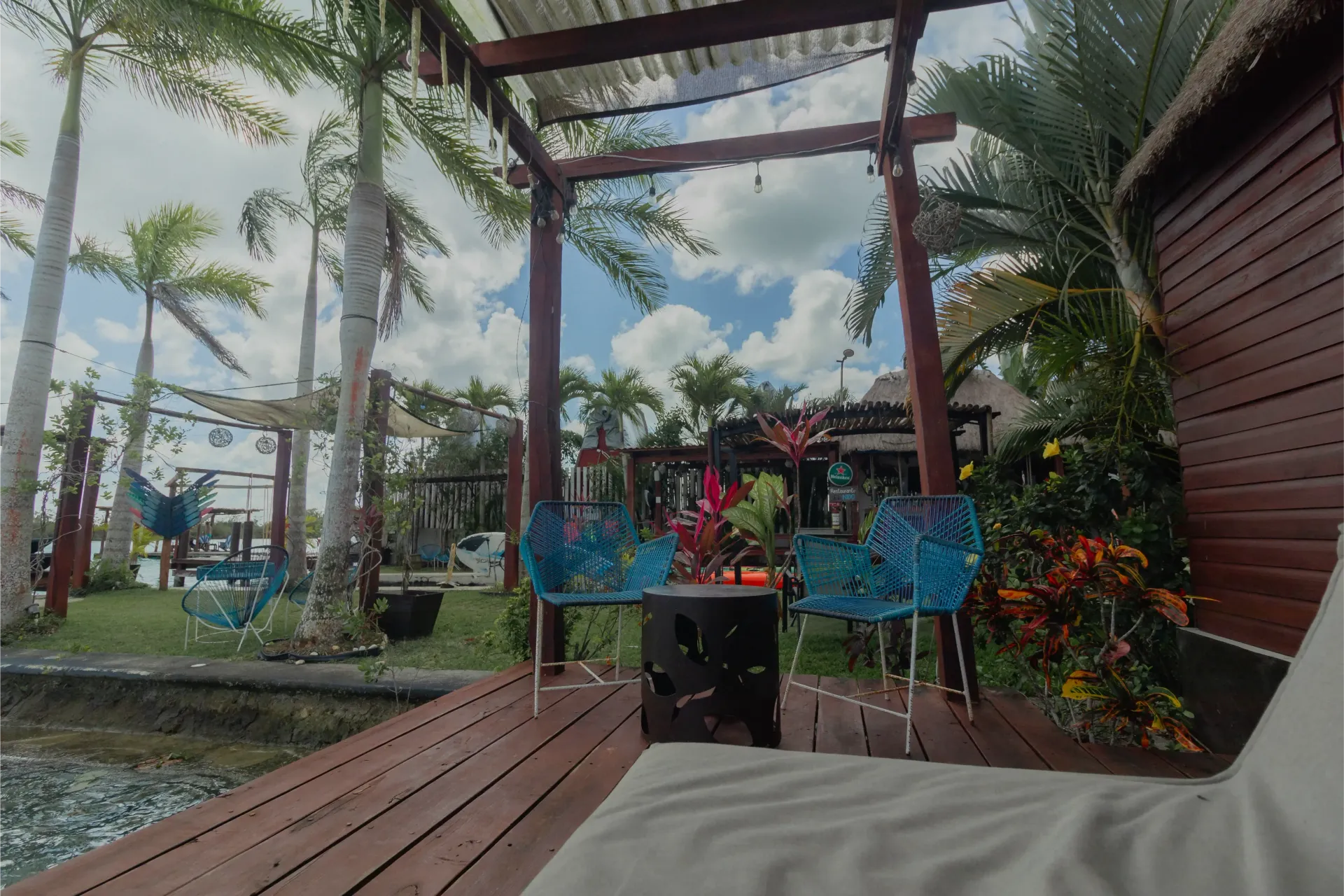 Patio exterior con terraza de madera, sillas azules, plantas tropicales y palmeras bajo un cielo nublado.