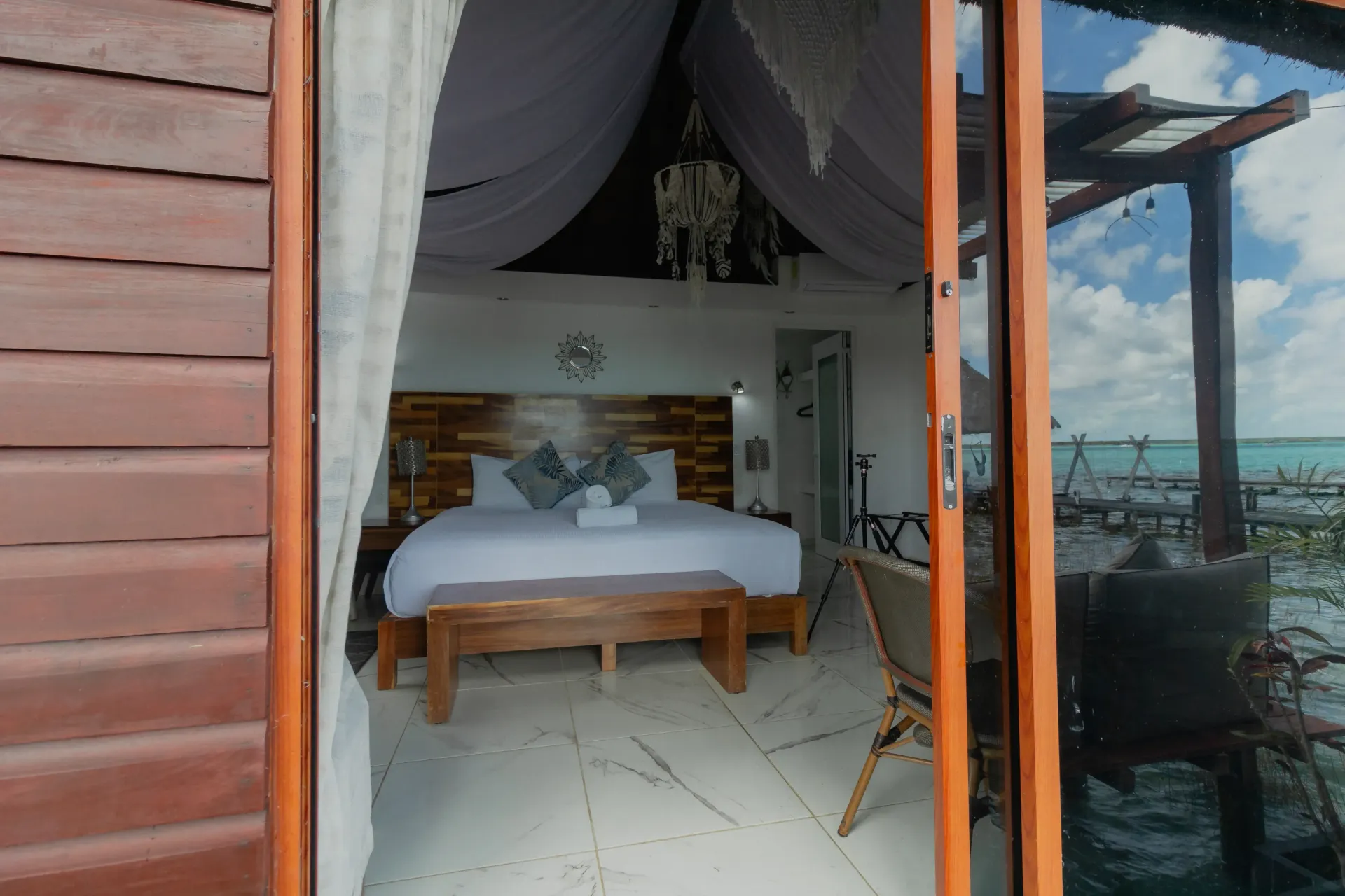 Habitación acogedora con cama blanca, banco de madera y puertas abiertas con vistas al mar.