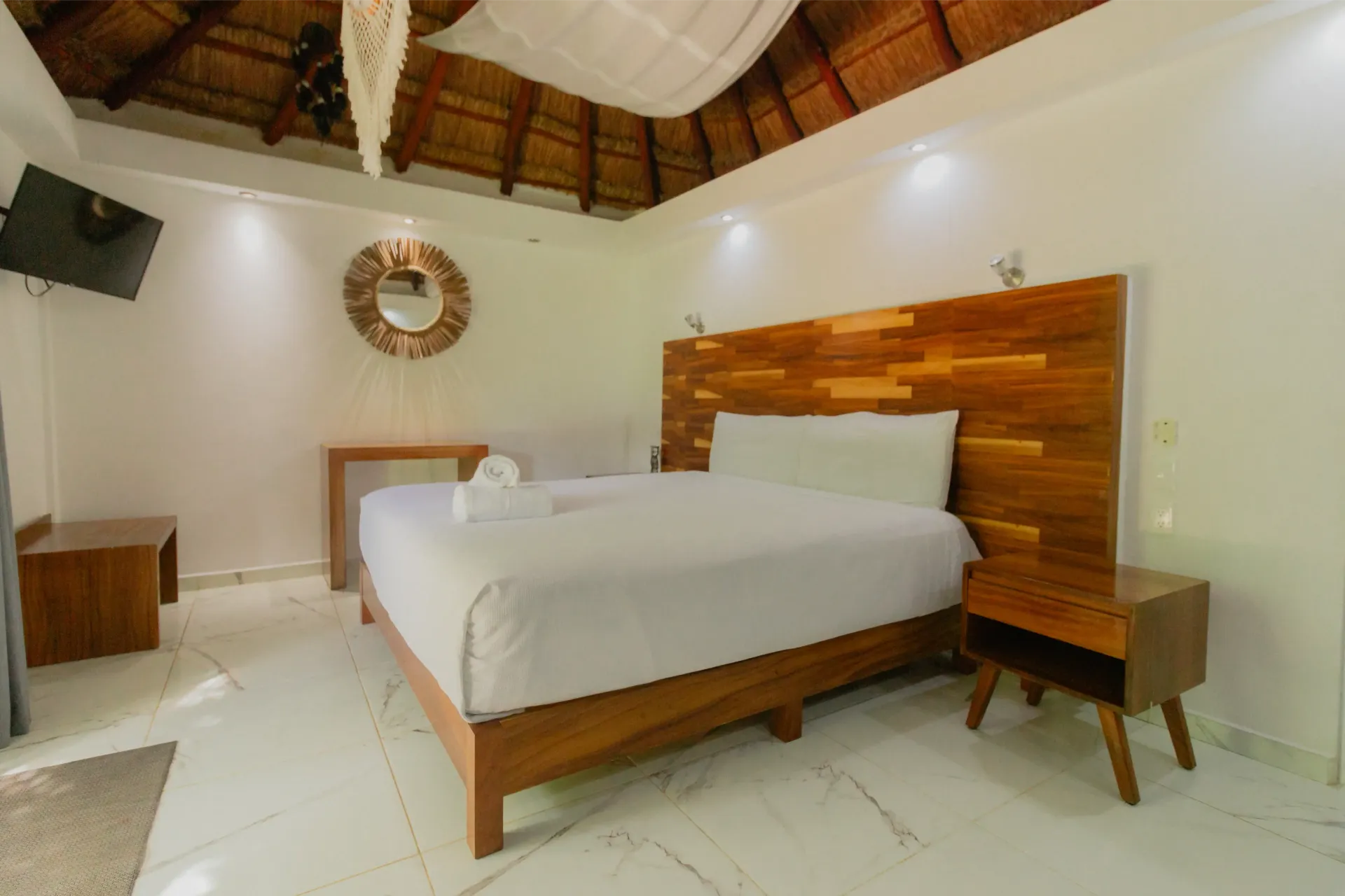 Habitación luminosa con cama de madera, ropa de cama blanca y techo de paja en un interior rústico.