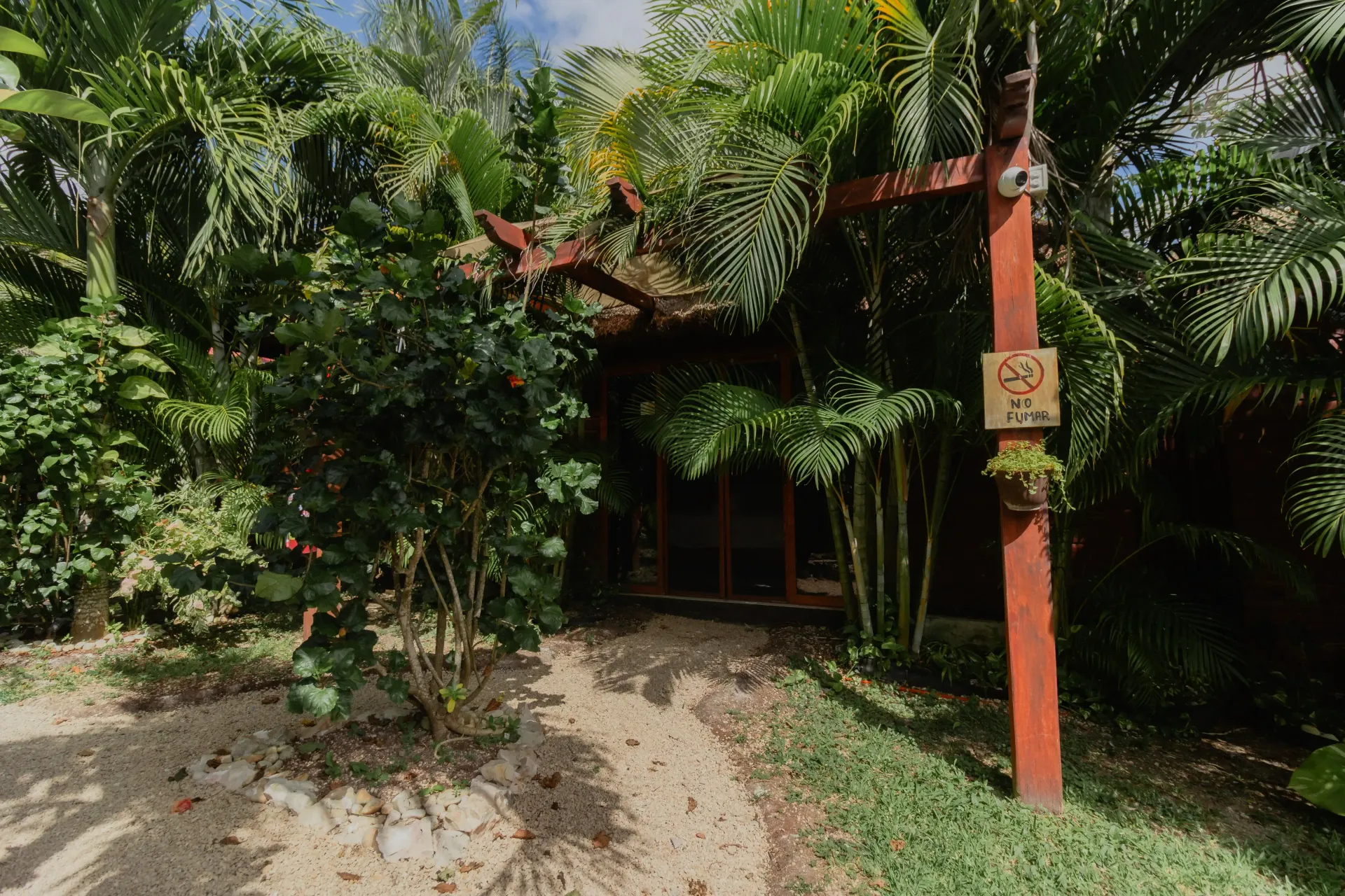 Entrada a un jardín tropical con vegetación densa, un poste rojo y un sendero arenoso que conduce a una puerta sombreada.
