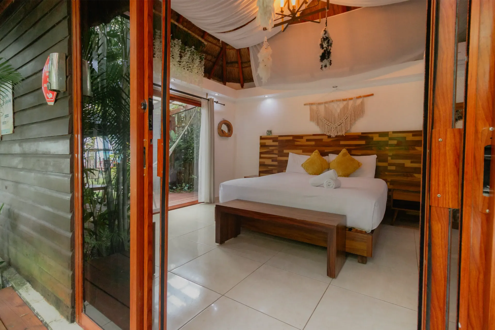 Dormitorio abierto con cama blanca, cabecero de madera, puertas correderas de cristal y decoración tropical en el techo.