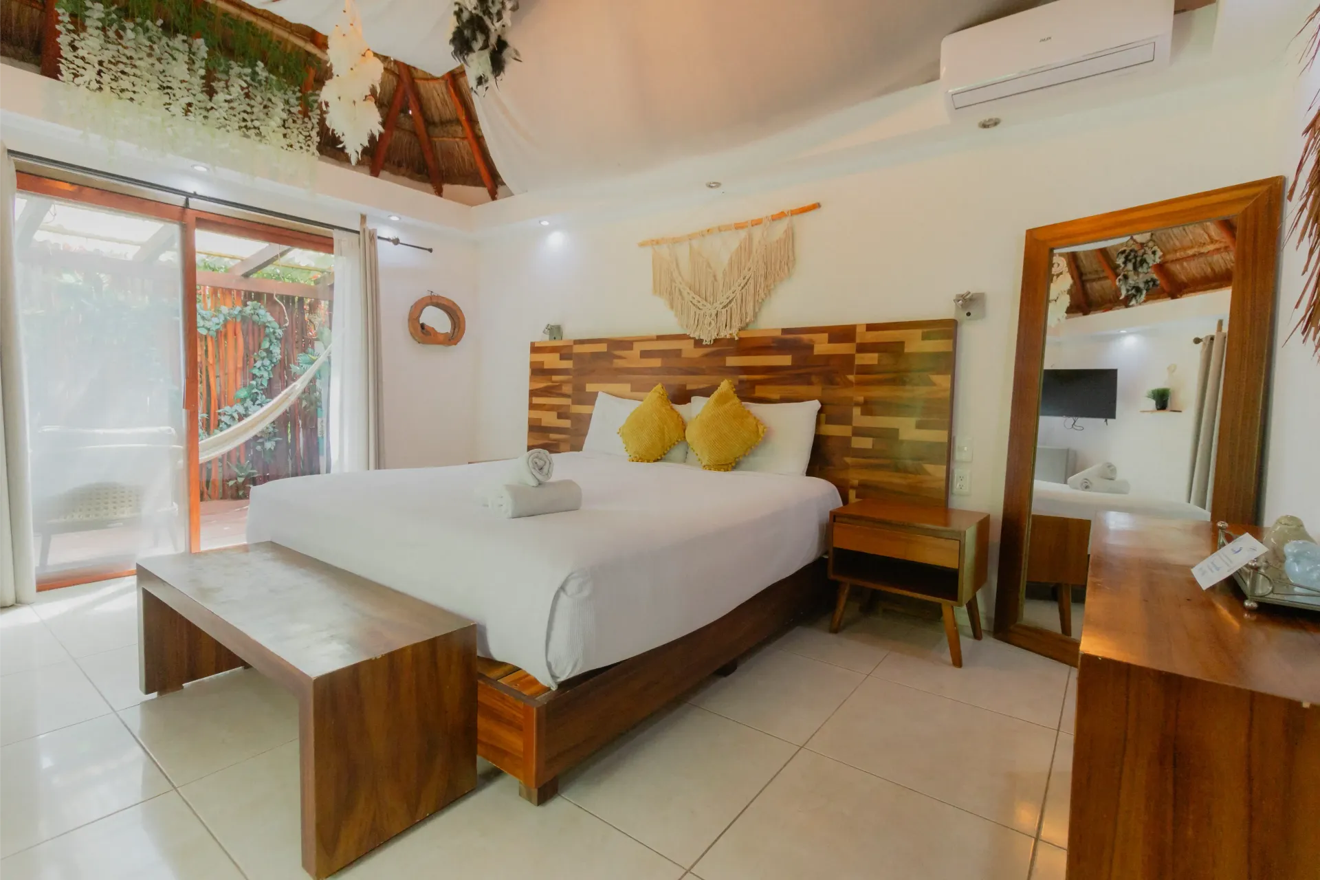 Habitación de hotel luminosa con cama blanca, muebles de madera, espejo y puerta corredera de cristal.