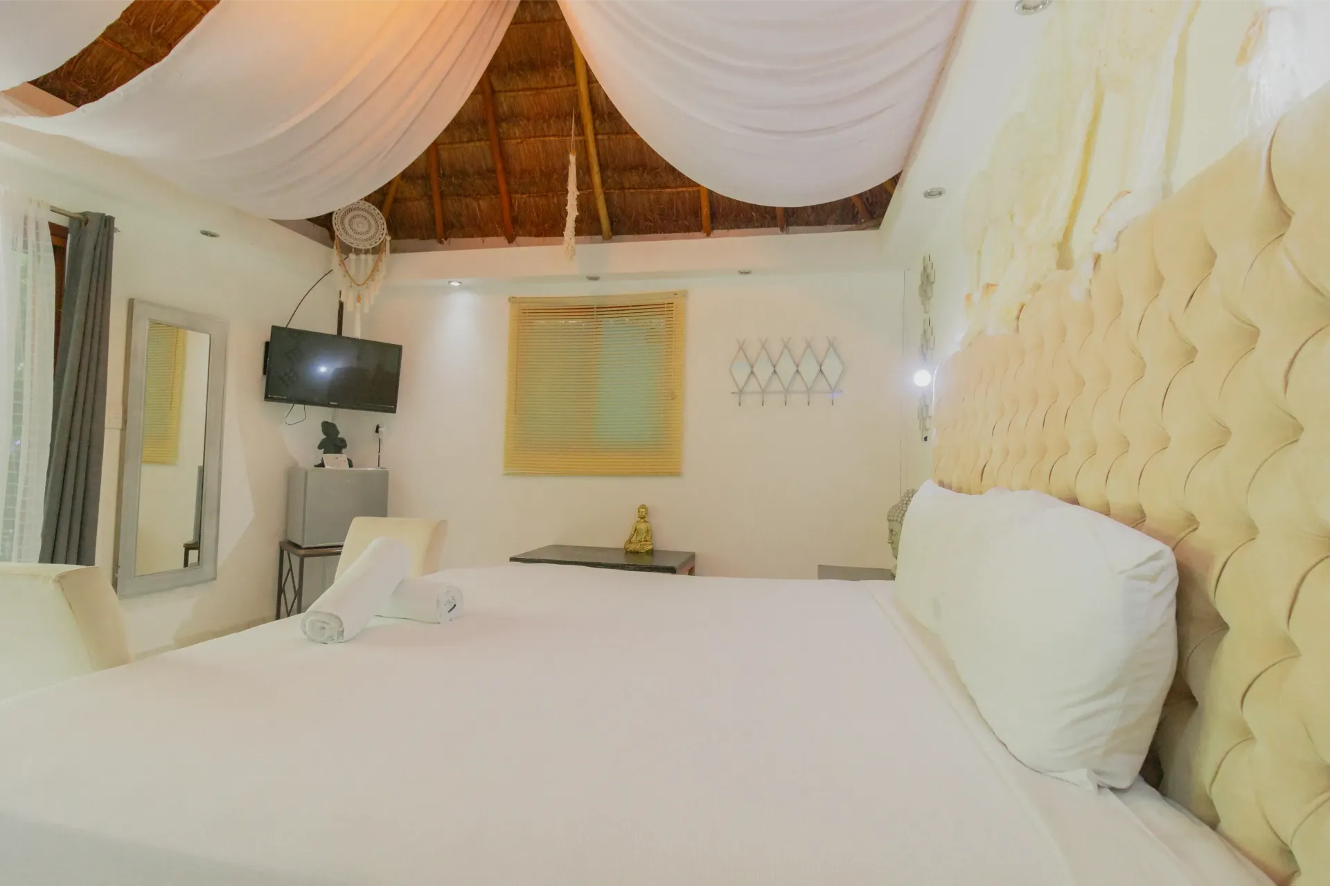 Elegante dormitorio con ropa de cama blanca, cabecero capitoné, dosel con cortinas y televisor montado en la pared.