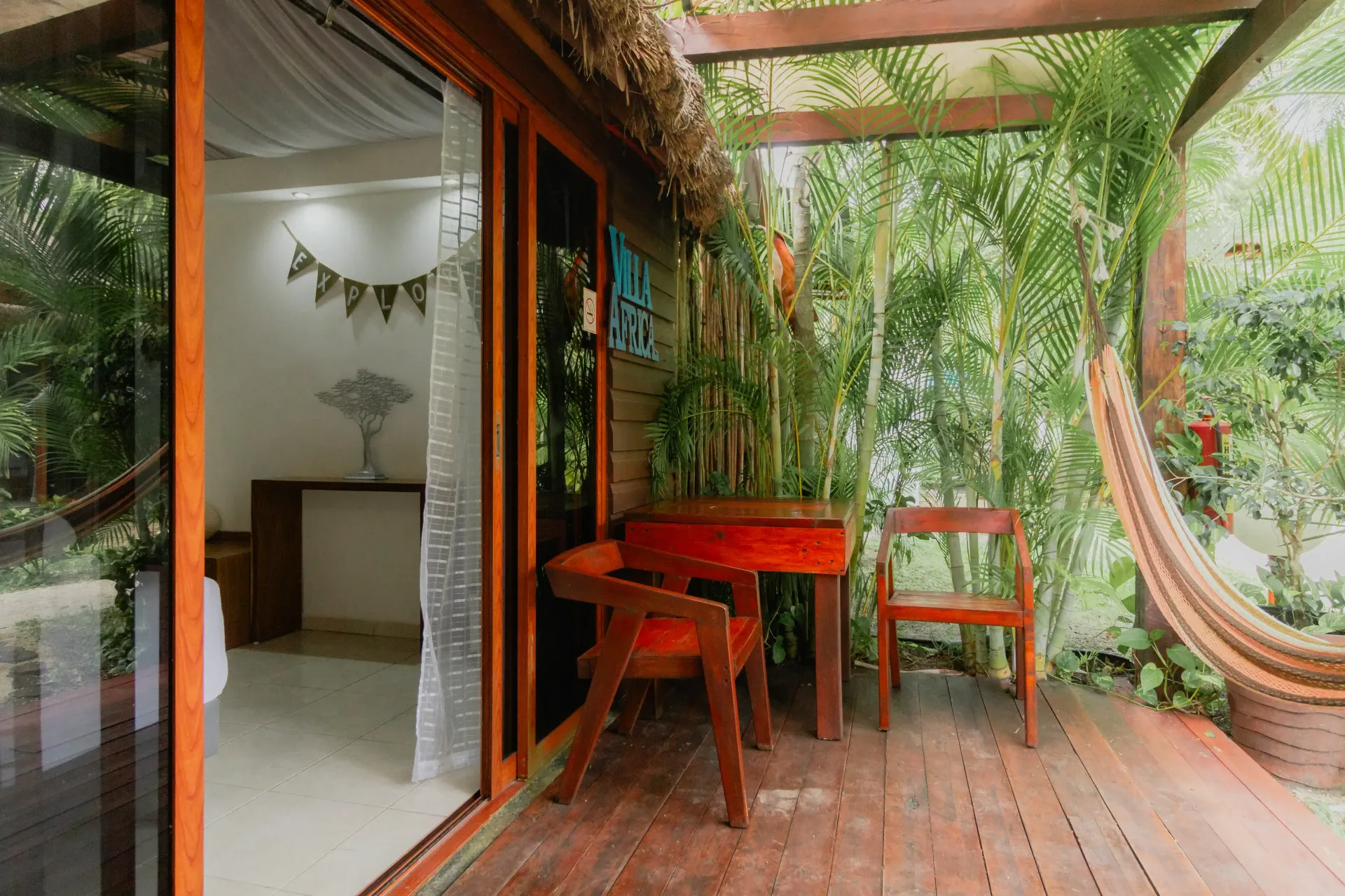 Patio tropical con mesa y sillas rojas, hamaca y una puerta corrediza abierta a un interior luminoso.