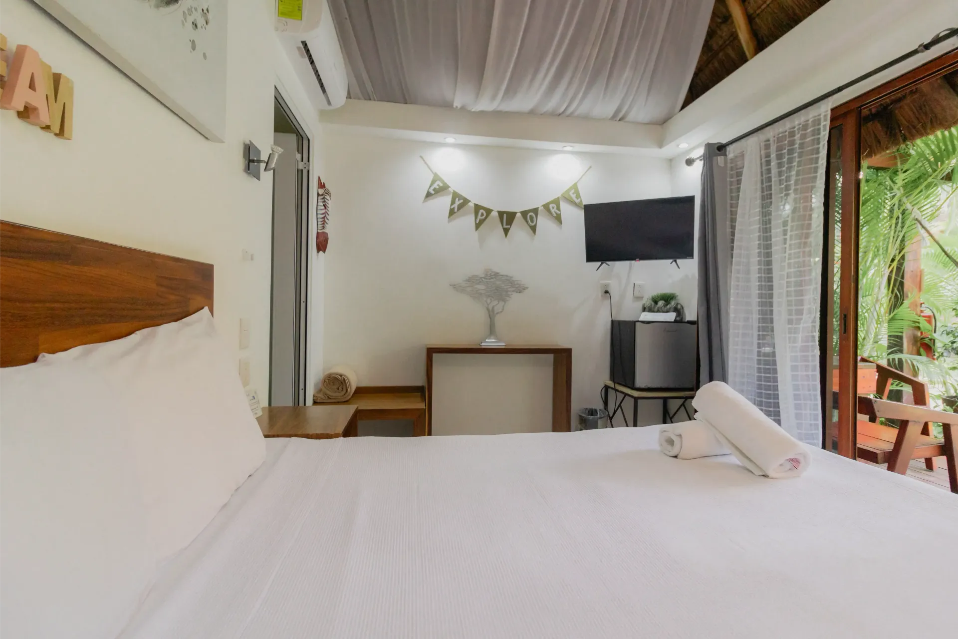 Habitación luminosa con cama blanca, dosel transparente, decoración de pared, escritorio, televisor y balcón abierto.